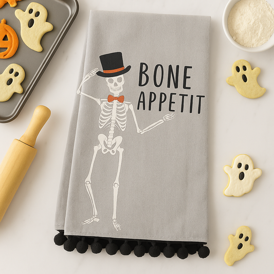 Gray towel with skeleton design and 'Bone Appetit' text, surrounded by Halloween-themed cookies and rolling pin.