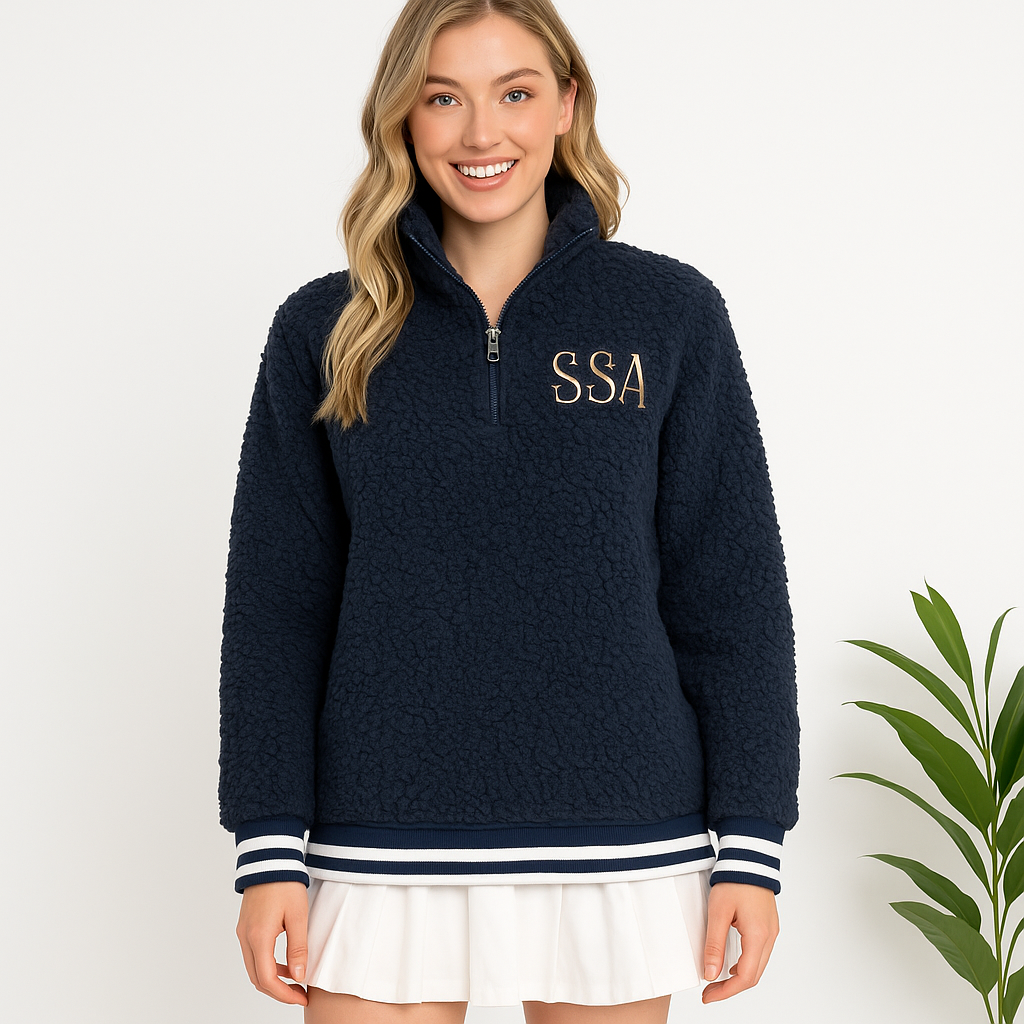 Young woman wearing navy sherpa pullover with SSA embroidered monogram and white pleated skirt, standing in front of white background with green plant